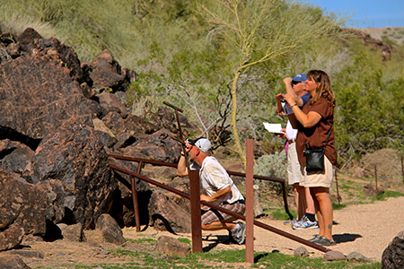 Three visitors shield their eyes from the sun as they look at petroglyphs