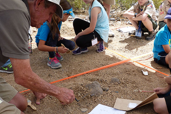 A volunteer demonstrates excavation techniques will children sit nearby